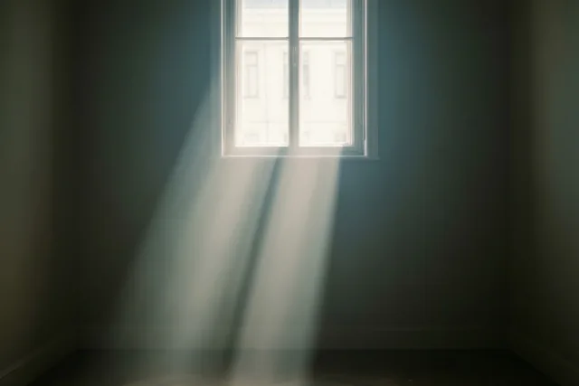 Een enkele zonnestraal valt door een hoog raam in een lege, schemerige kamer met donkere muren en een houten vloer.