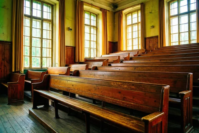 Een oude aula met houten banken, gele muren en hoge ramen ligt in warm zonlicht, met groen buiten de ramen.