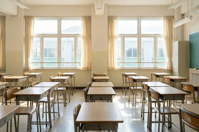 Een lege klas met beige muren, lichtgele gordijnen en rijen houten banken en stoelen baadt in zonlicht uit hoge ramen.