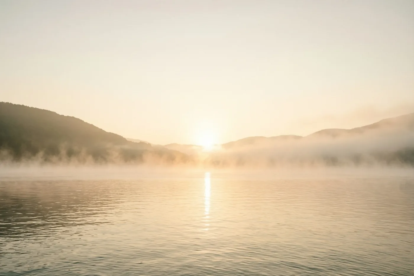De zon komt op boven rustig water met optrekkende mist en heuvels in de verte, in goudkleurige en lichte perziktinten.