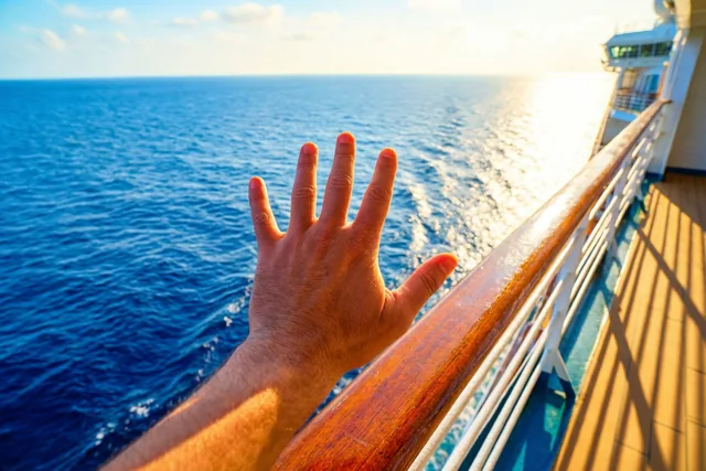 Een hand zwaait bij de houten reling van een cruiseschip, met een uitgestrekte blauwe zee onder een heldere hemel in warm zonlicht.