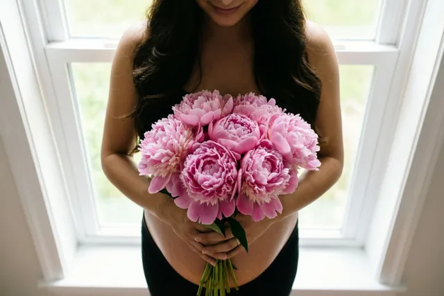 Een zwangere vrouw staat bij een helder raam met roze pioenen tegen haar buik, in zacht daglicht en met roze, zwart en wit als hoofdkleuren.