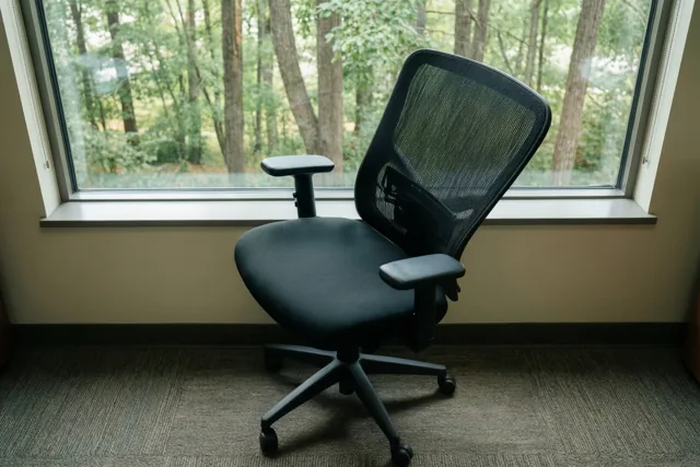 Een lege zwarte ergonomische bureaustoel staat op een tapijt met een patroon, schuin naar een groot raam met uitzicht op groene bomen in daglicht.