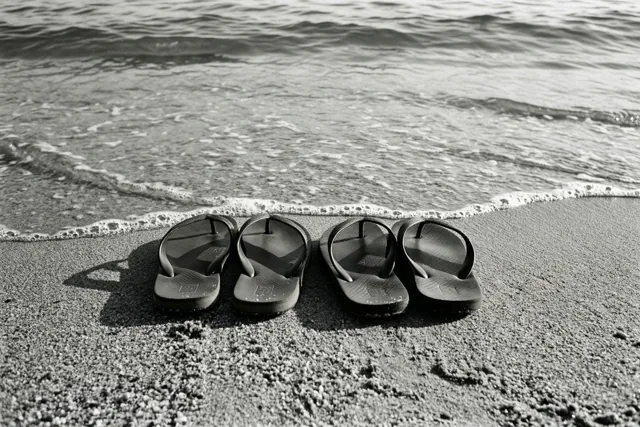 Twee paar zwarte teenslippers liggen naast elkaar op nat zand bij zachte golven aan de waterlijn, op een zwart-witfoto.