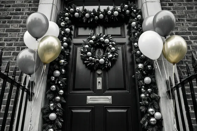 Een zwarte voordeur in een bakstenen muur is versierd met een slinger en ballonnen in zwart, wit en goud, met een zwart metalen hek ervoor.